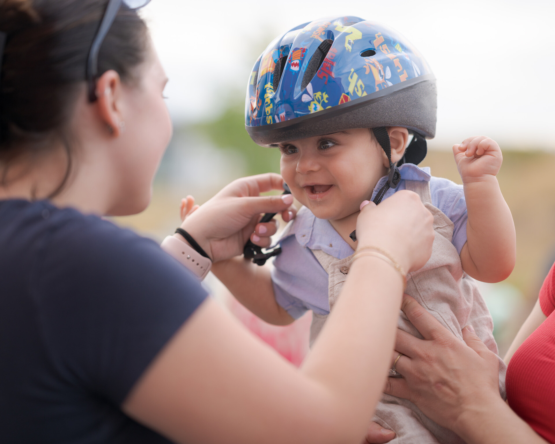 An adult fastens a blue helmet with cartoon designs on a smiling baby, while another supports the child, promoting CPFR driver and pedestrian safety.