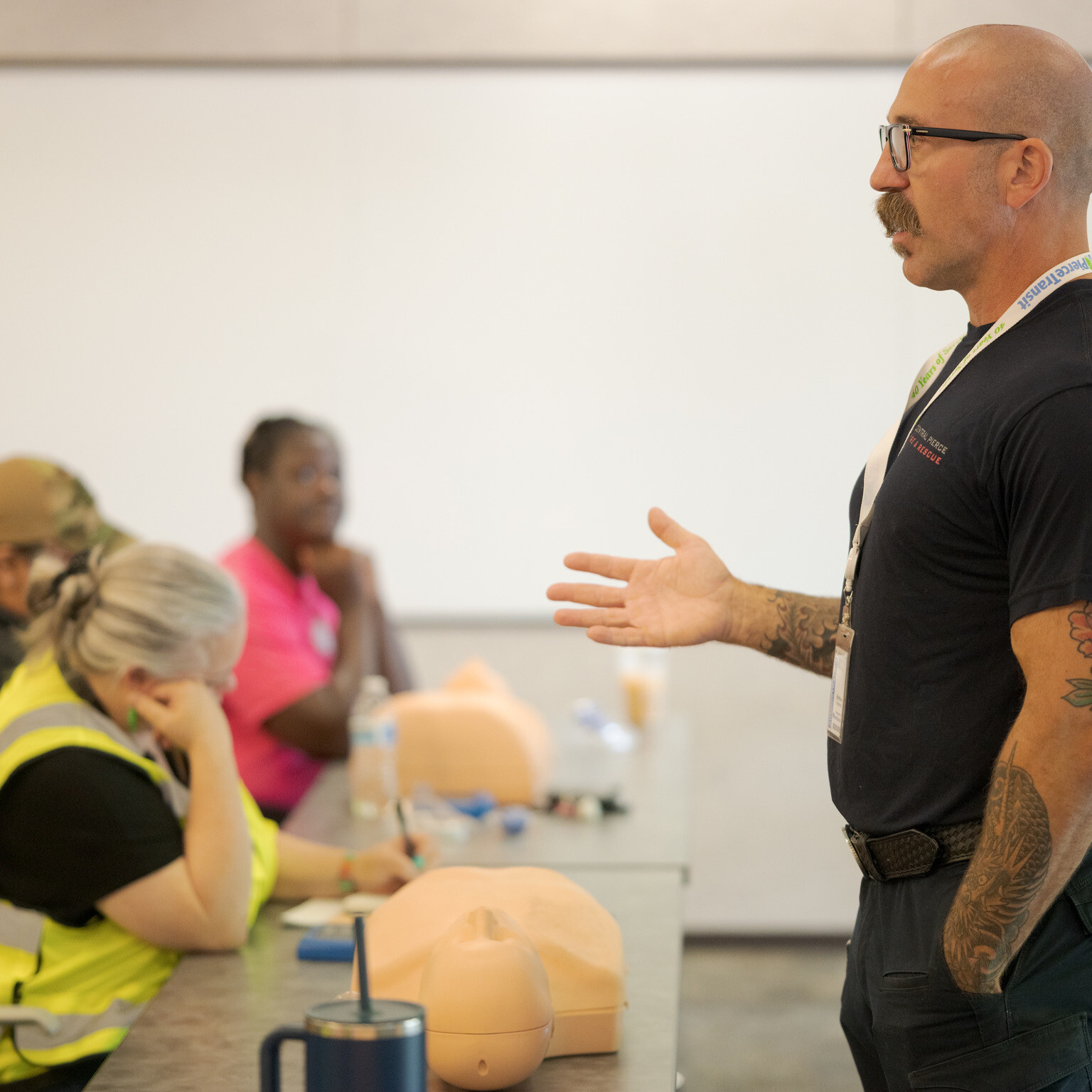 A man stands and speaks to a seated group during a CPR training session, with practice mannequins on the table in front of participants.