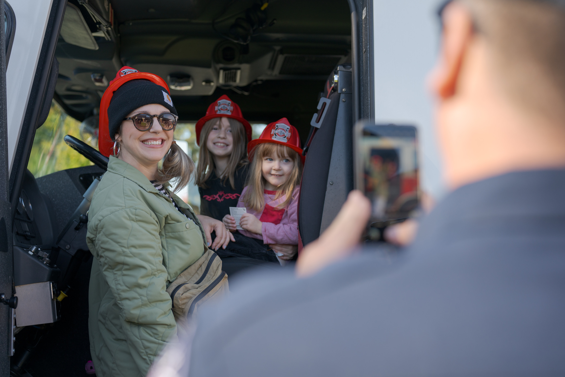 A woman and two young girls wearing red plastic fire helmets sit inside a vehicle while a person takes their photo with a smartphone.