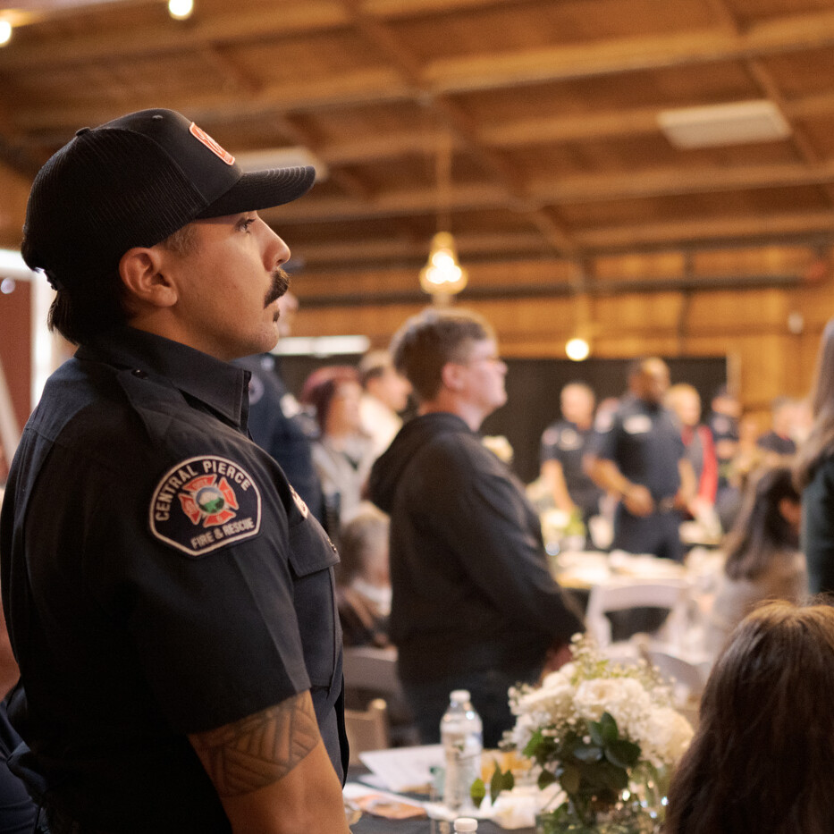 A group of people, including uniformed firefighters, stand in a wooden hall with tables, floral arrangements, and soft lighting.
