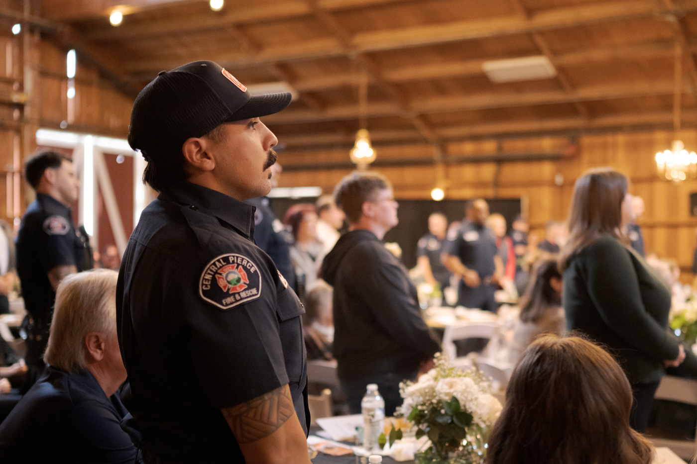 A group of people, including uniformed firefighters, stand in a wooden hall with tables, floral arrangements, and soft lighting.