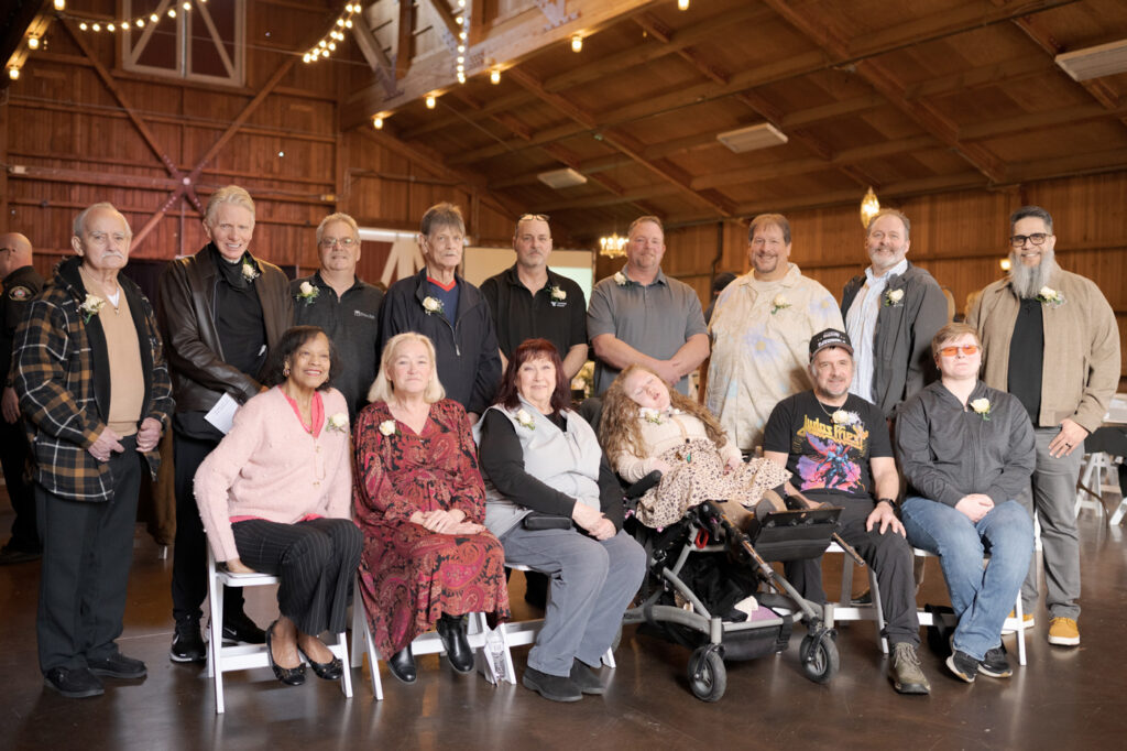 A group of fourteen adults, some seated and some standing, pose for a group photo in a wooden hall decorated with string lights.
