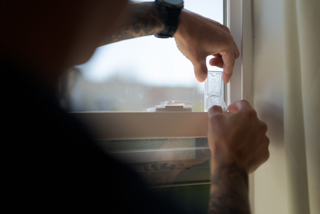 Person installs a plastic device on a window frame with both hands. The window is partially open, and the background is out of focus.