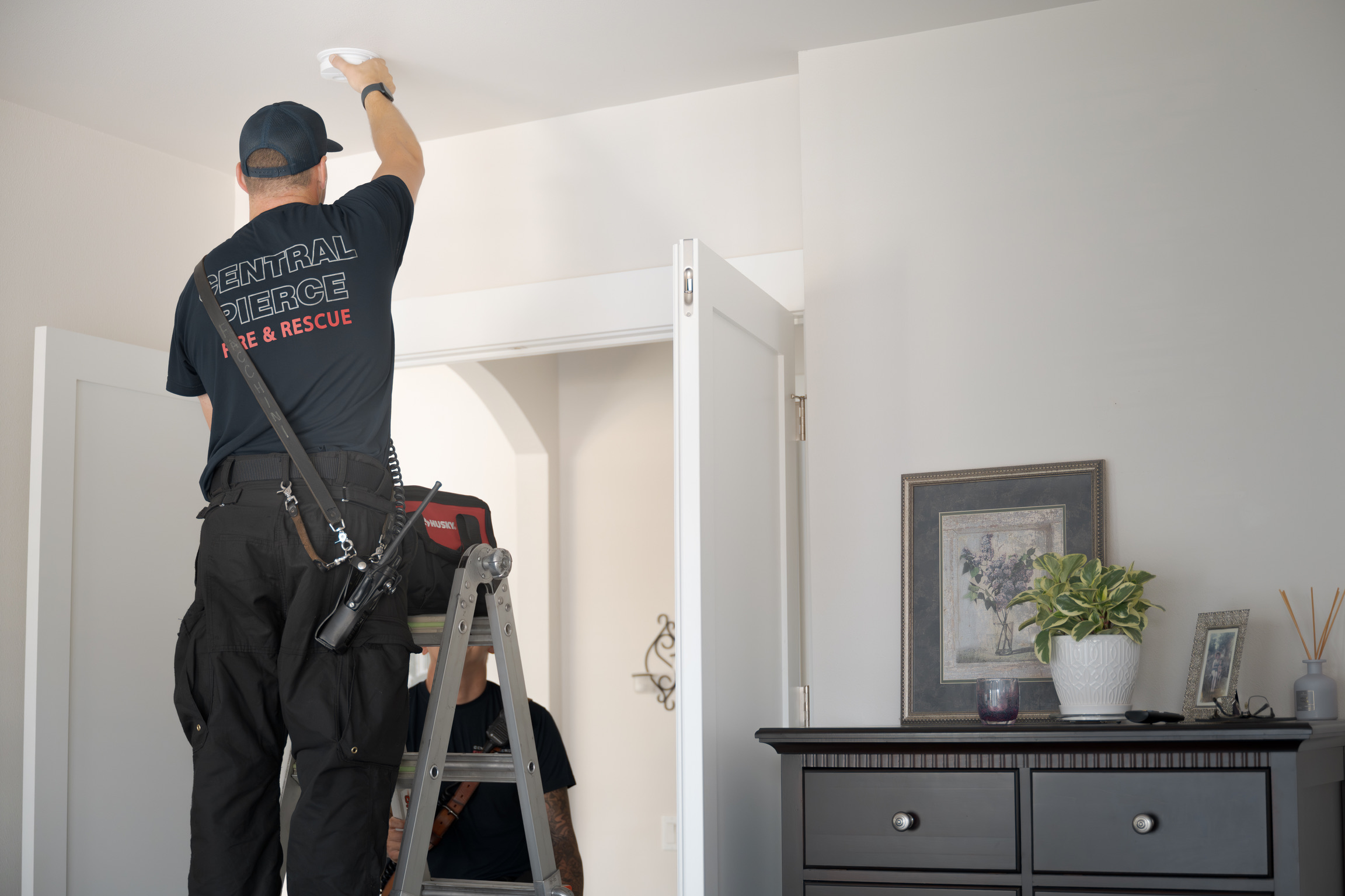 A firefighter stands on a ladder inspecting or installing a smoke detector on the ceiling inside a home.