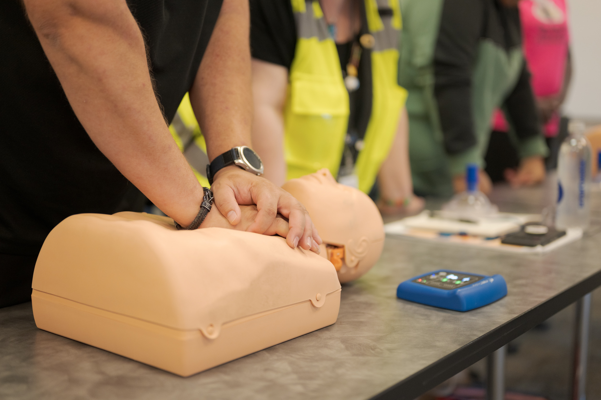 Person practicing CPR chest compressions on a training mannequin, with others nearby and a defibrillator device on the table.