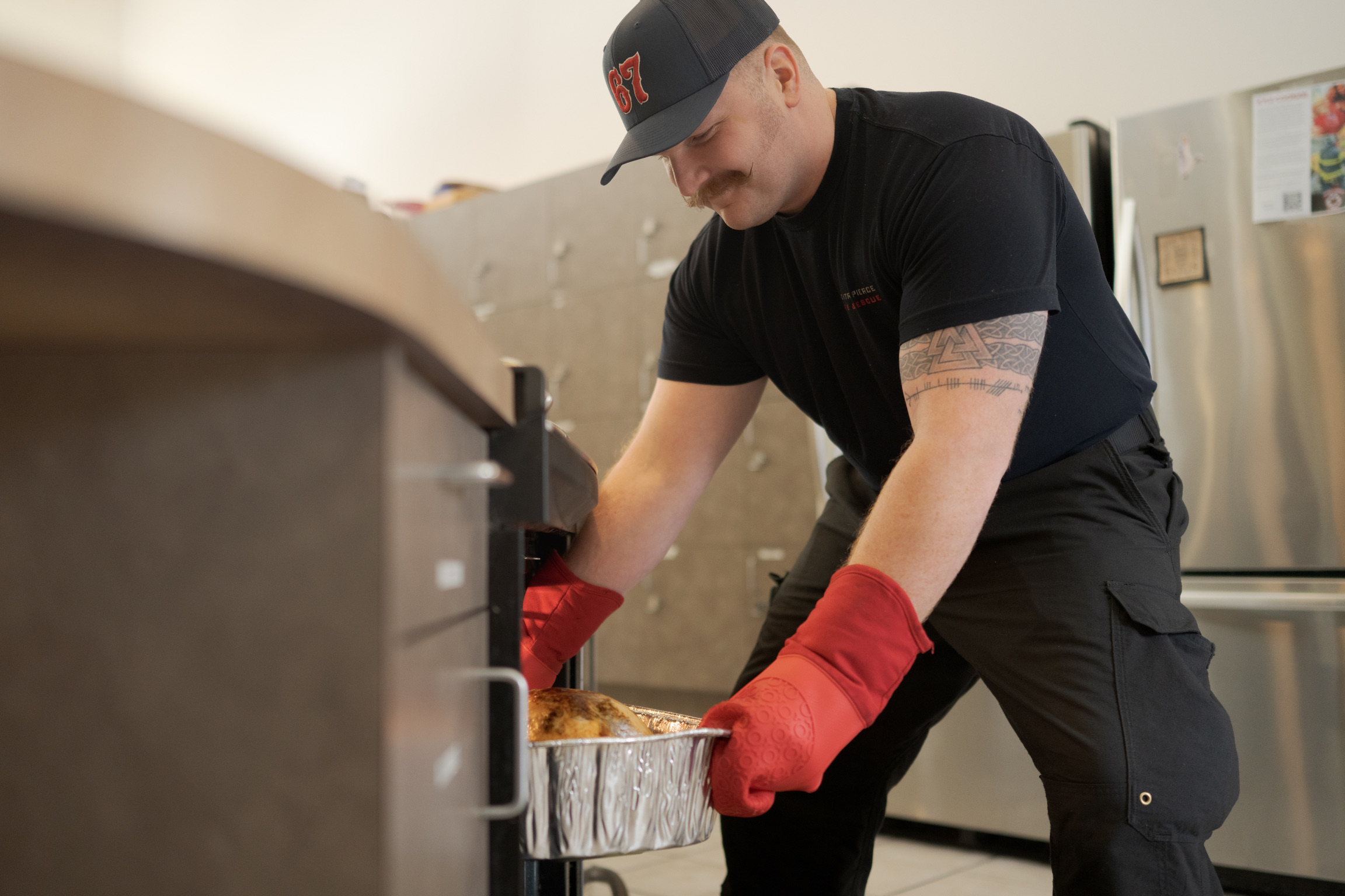 A man wearing red oven mitts places a tray of food into an oven in a kitchen.