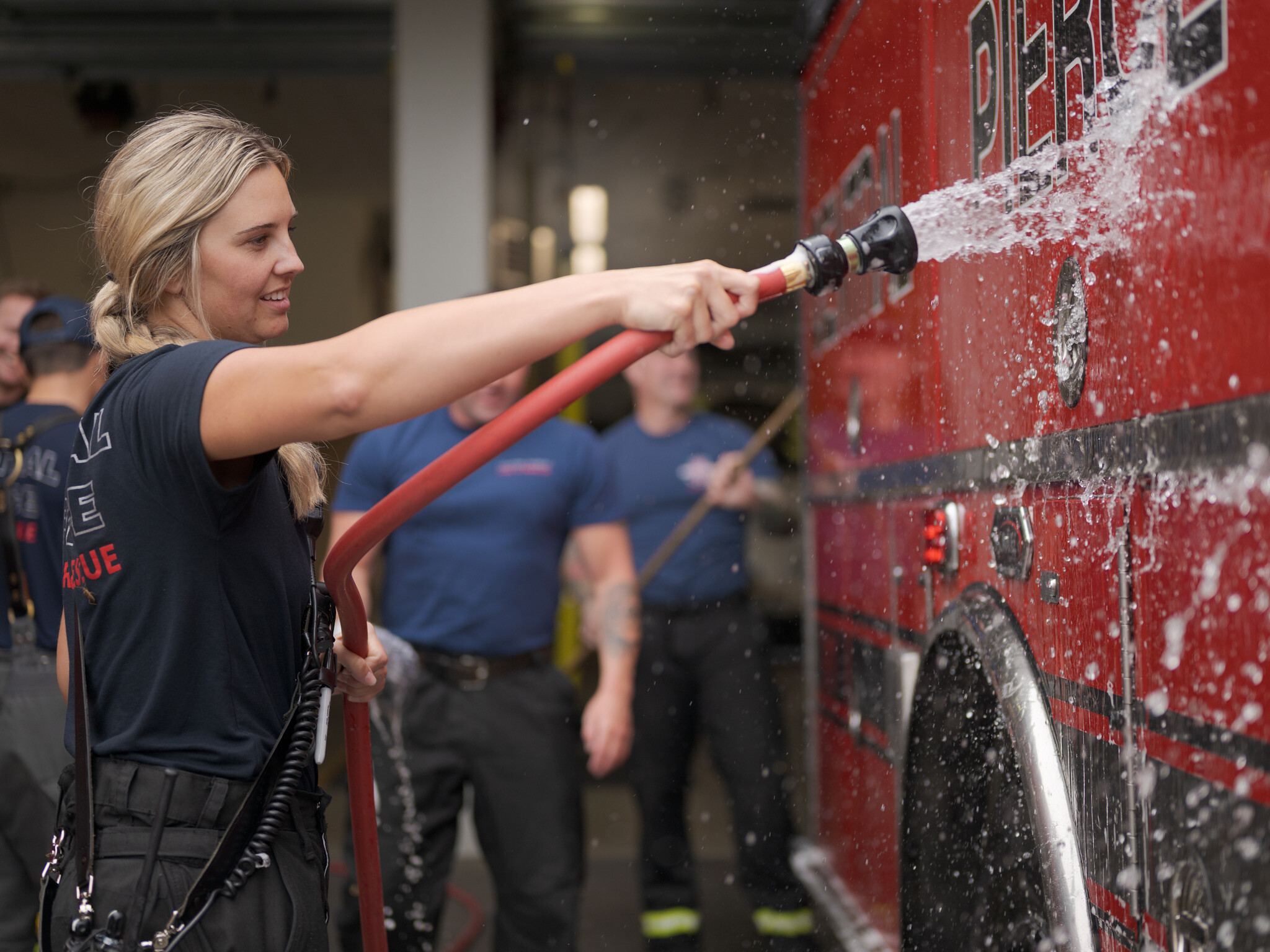 A firefighter uses a hose to wash a red fire truck while other firefighters stand in the background at Central Pierce Fire & Rescue, where Ride-Along Requests are welcome for those interested in experiencing a day with the crew.