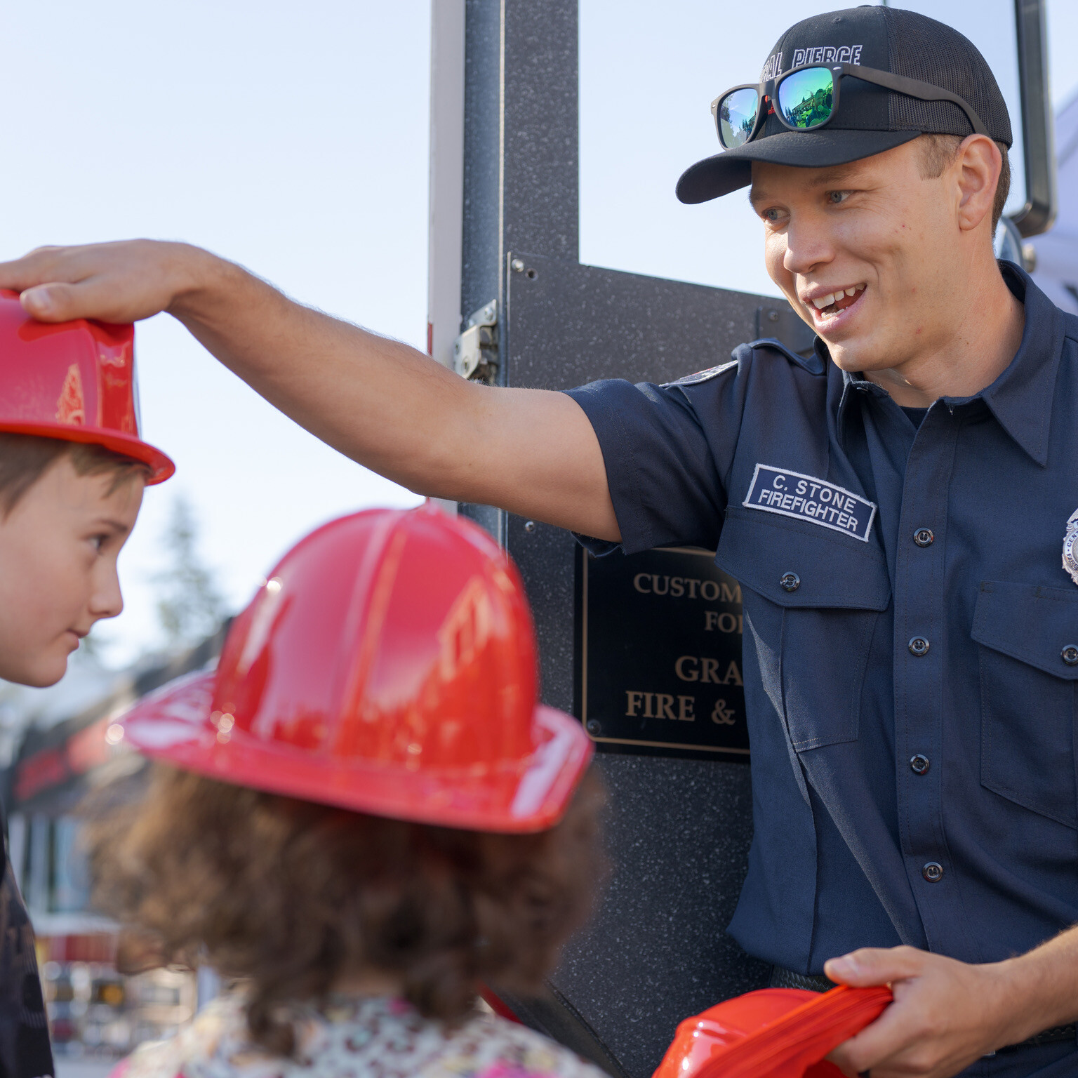 A firefighter places a red helmet on a boy&rsquo;s head while talking to two children at an outdoor community event.