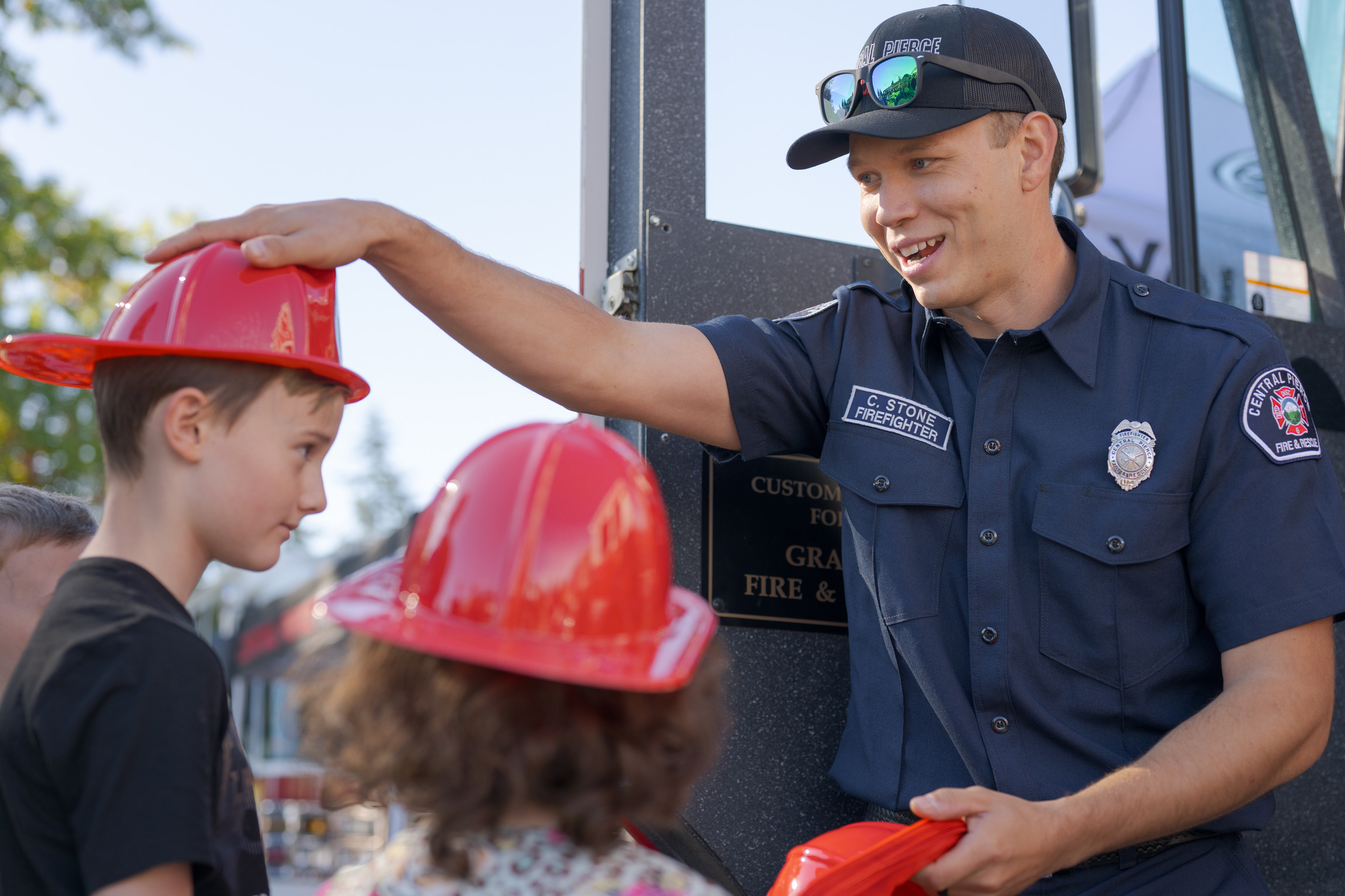 A firefighter places a red helmet on a boy’s head while talking to two children at an outdoor community event.