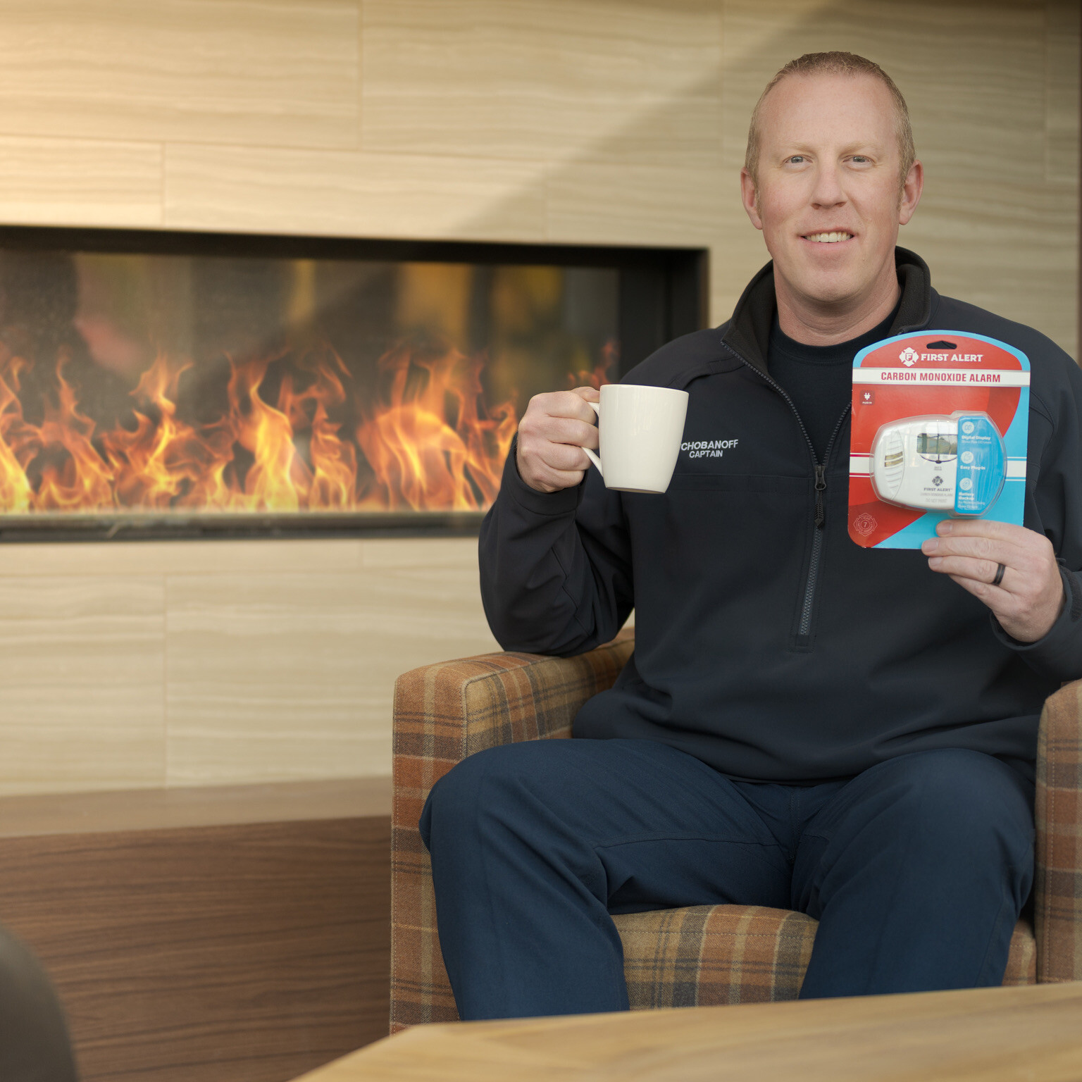 A man sits in a chair by a modern fireplace, holding a white mug in one hand and a packaged smoke and carbon monoxide alarm in the other.