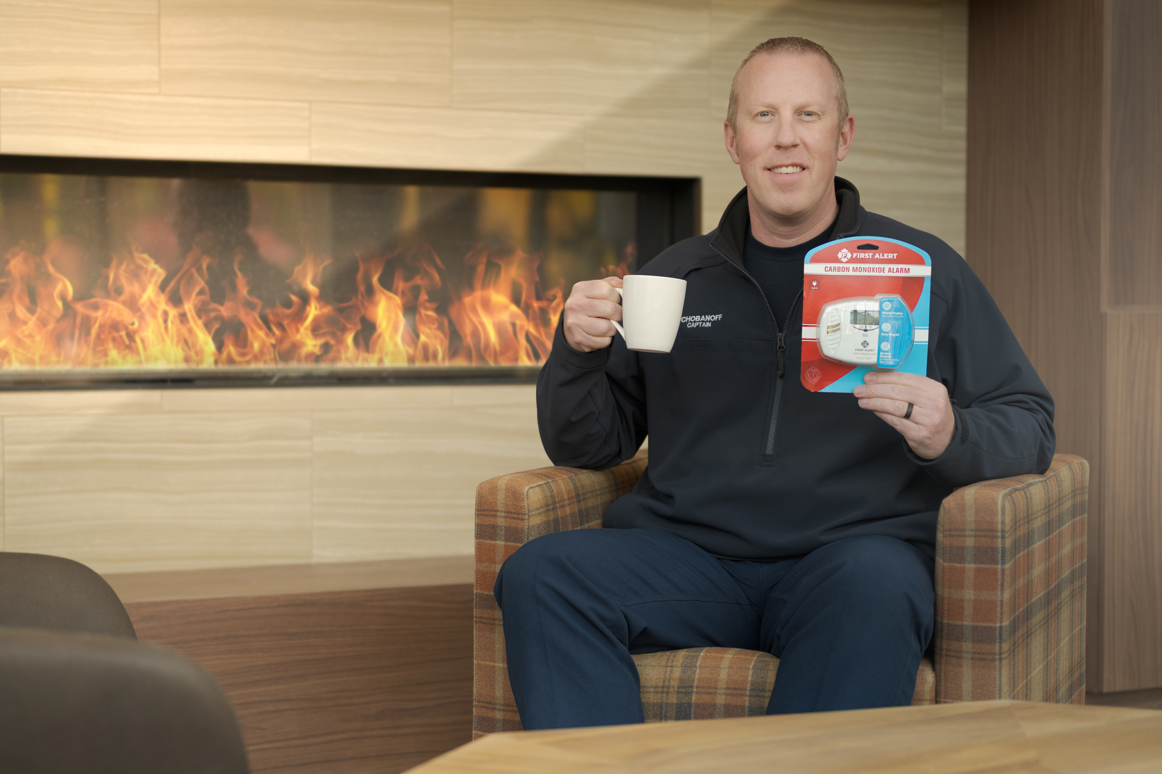 A man sits in a chair by a modern fireplace, holding a white mug in one hand and a packaged smoke and carbon monoxide alarm in the other.