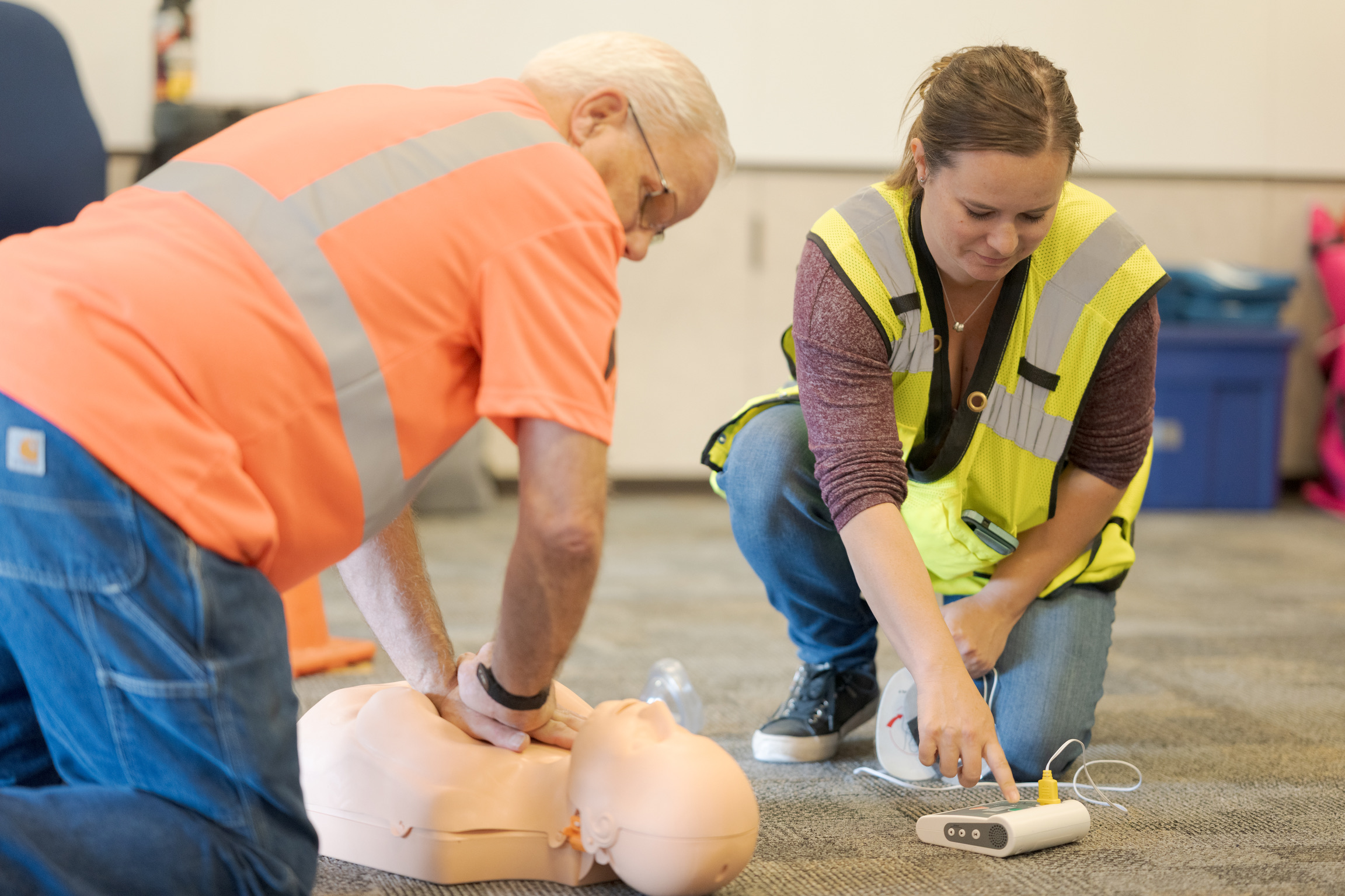 Two people in safety vests practice CPR and use a training defibrillator on a mannequin in a classroom setting.