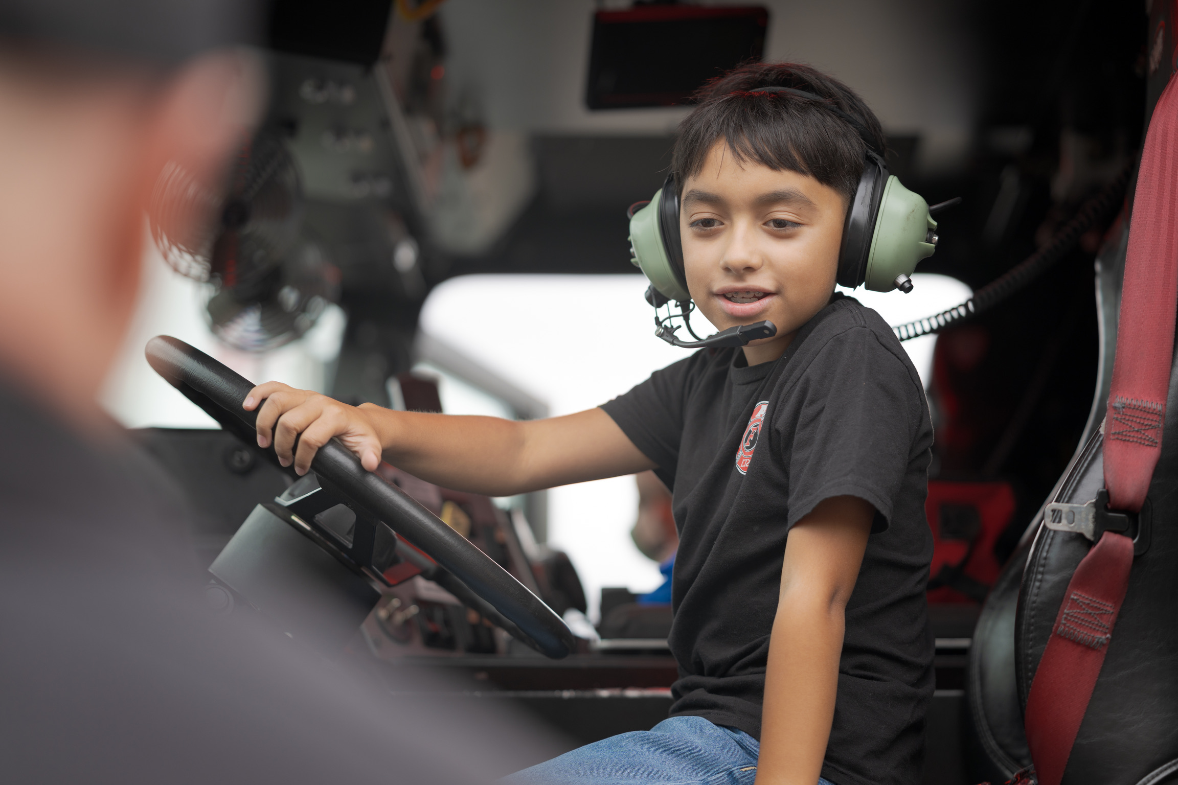 A boy wearing a headset sits in the driver's seat of a large vehicle, holding the steering wheel and looking ahead.