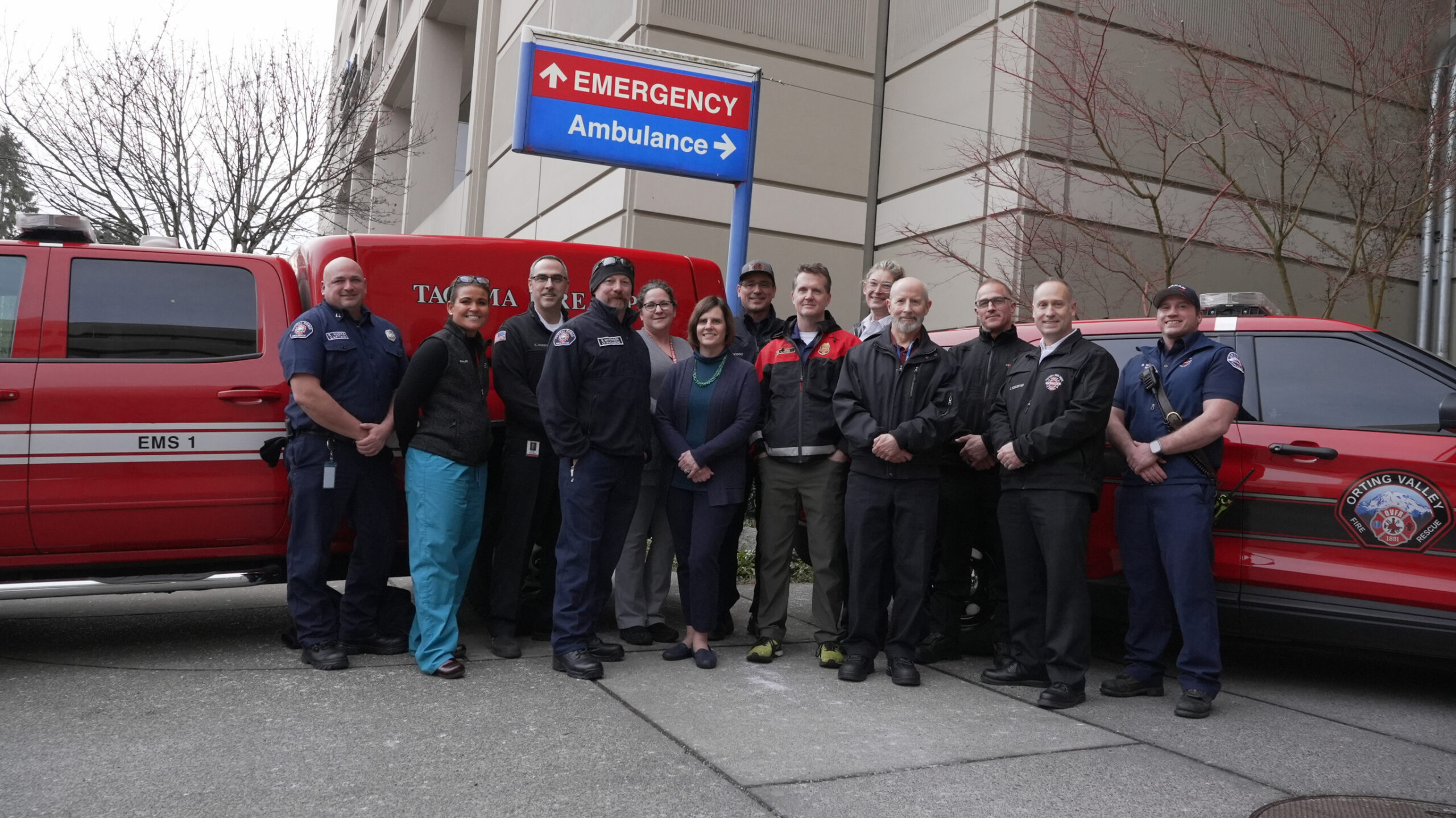 A group of emergency personnel from Central Pierce Fire & Rescue and hospital staff stand together outside near red emergency vehicles, an emergency room sign, and carbon monoxide alarms.