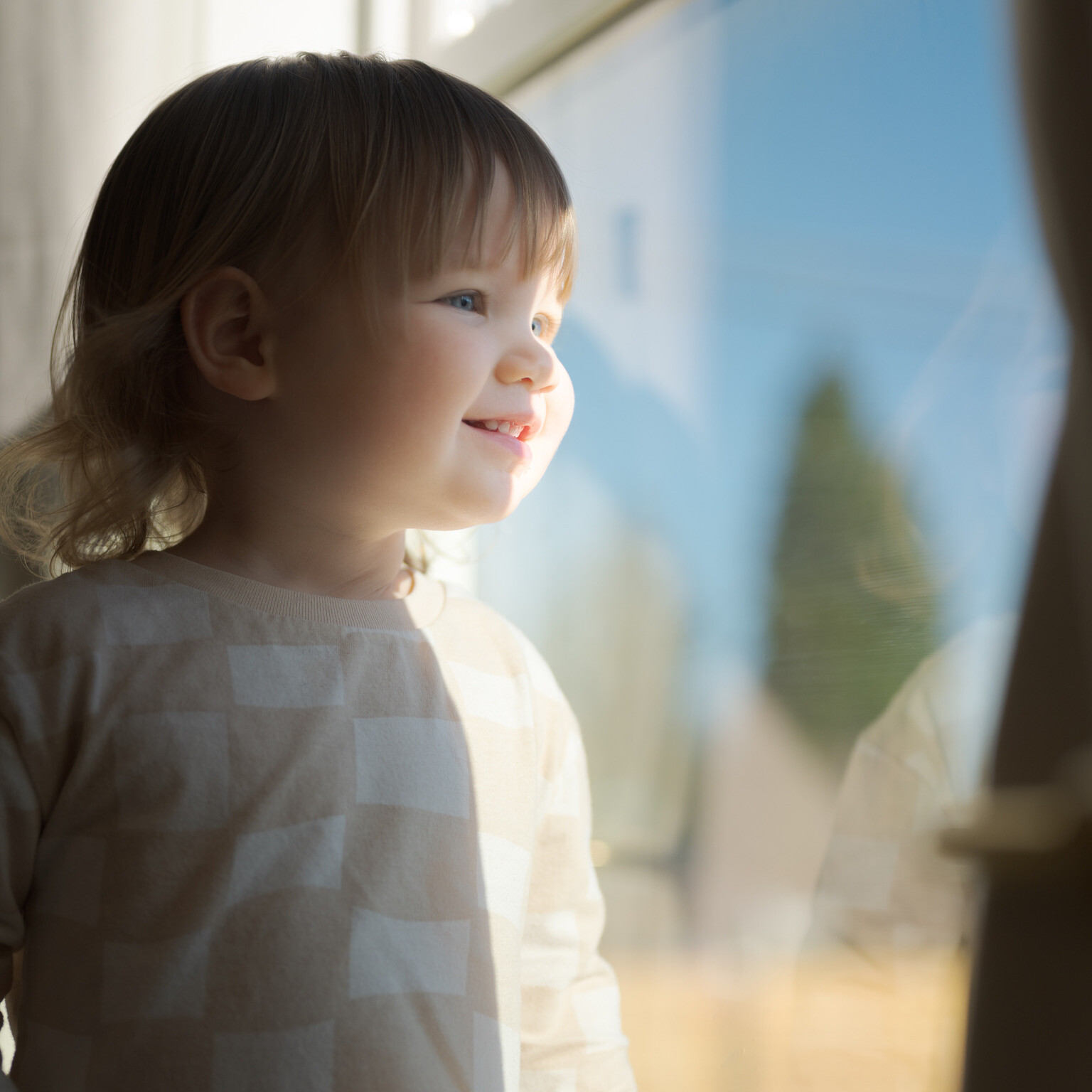 A young child with light hair smiles while looking out a sunlit window, wearing a patterned long-sleeve shirt.