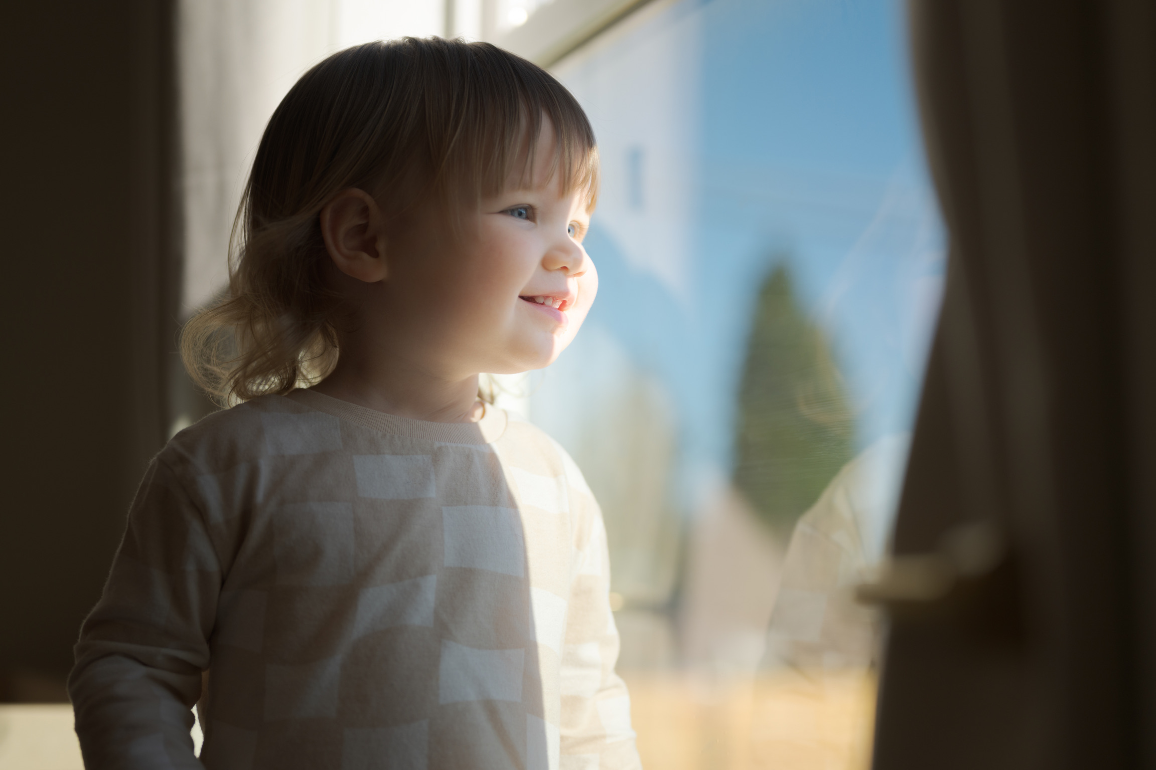 A young child with light hair smiles while looking out a sunlit window, wearing a patterned long-sleeve shirt.