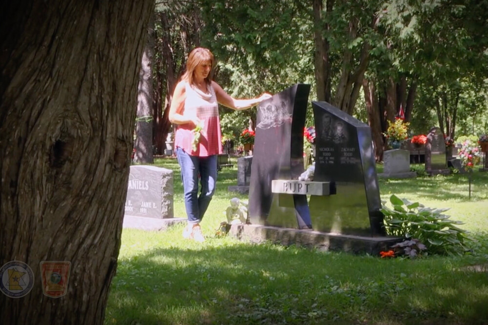 A woman stands beside and touches a gravestone in a sunlit cemetery surrounded by trees and other headstones.
