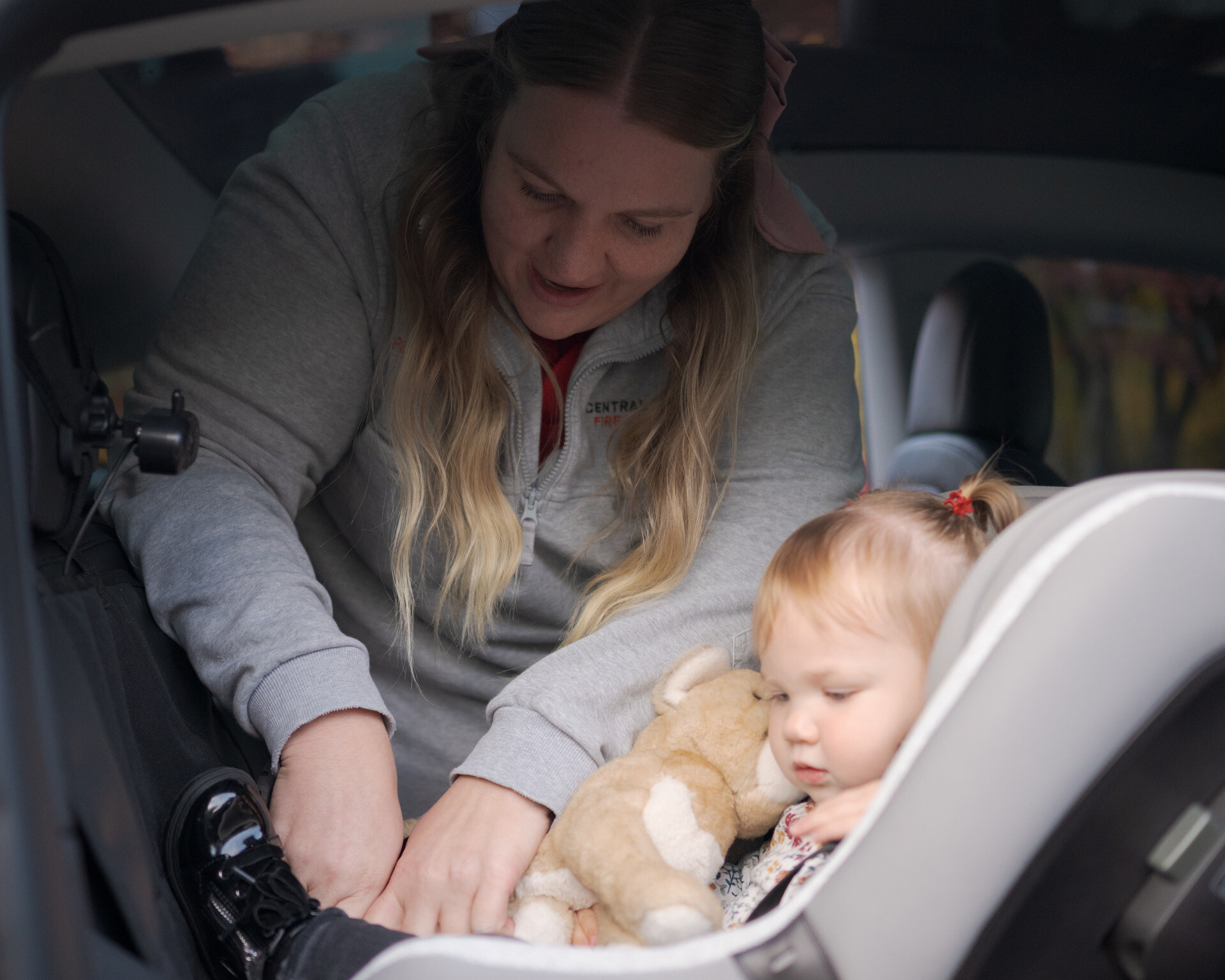An adult secures a young child into a car seat in the back of a vehicle. The child is holding a stuffed animal and appears calm.