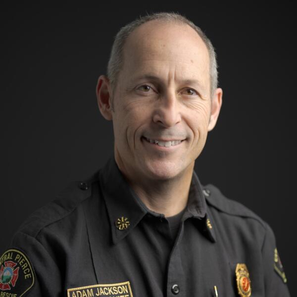 A middle-aged man in a dark uniform with badges and an embroidered name tag that reads 