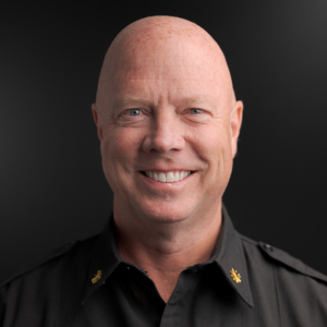 A bald man in a dark uniform shirt with gold insignia smiles at the camera in front of a dark background.