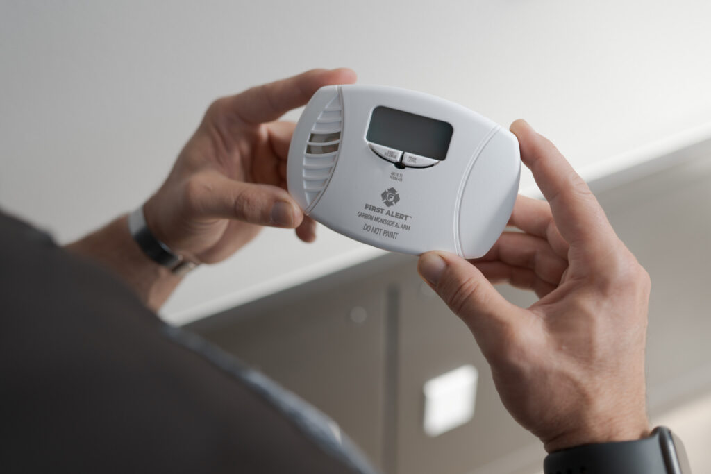 Person holds a First Alert carbon monoxide detector with a digital display, preparing to install or test it indoors.