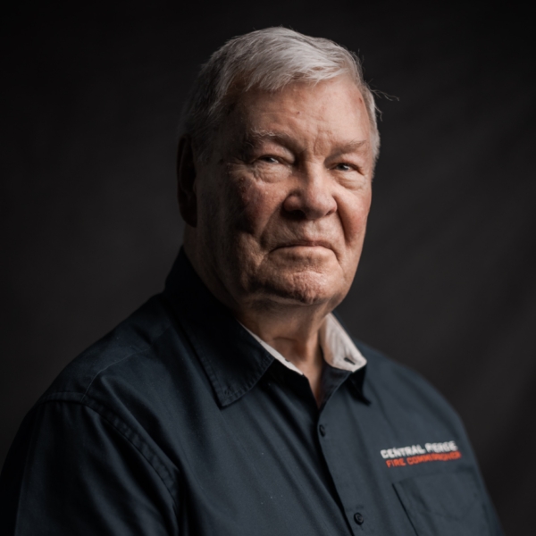 Older man with gray hair wearing a dark button-up shirt, facing slightly to the side, photographed against a dark background.