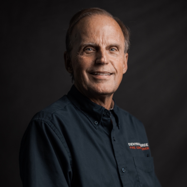 An older man with short hair smiles at the camera, wearing a dark button-up shirt with embroidered text, against a dark background.
