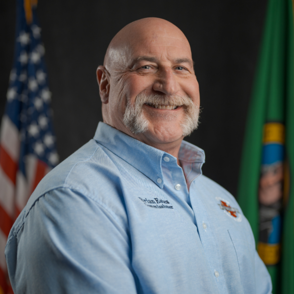 A man with a bald head and mustache smiles while standing in front of American and Washington state flags, wearing a light blue collared shirt with embroidered text.