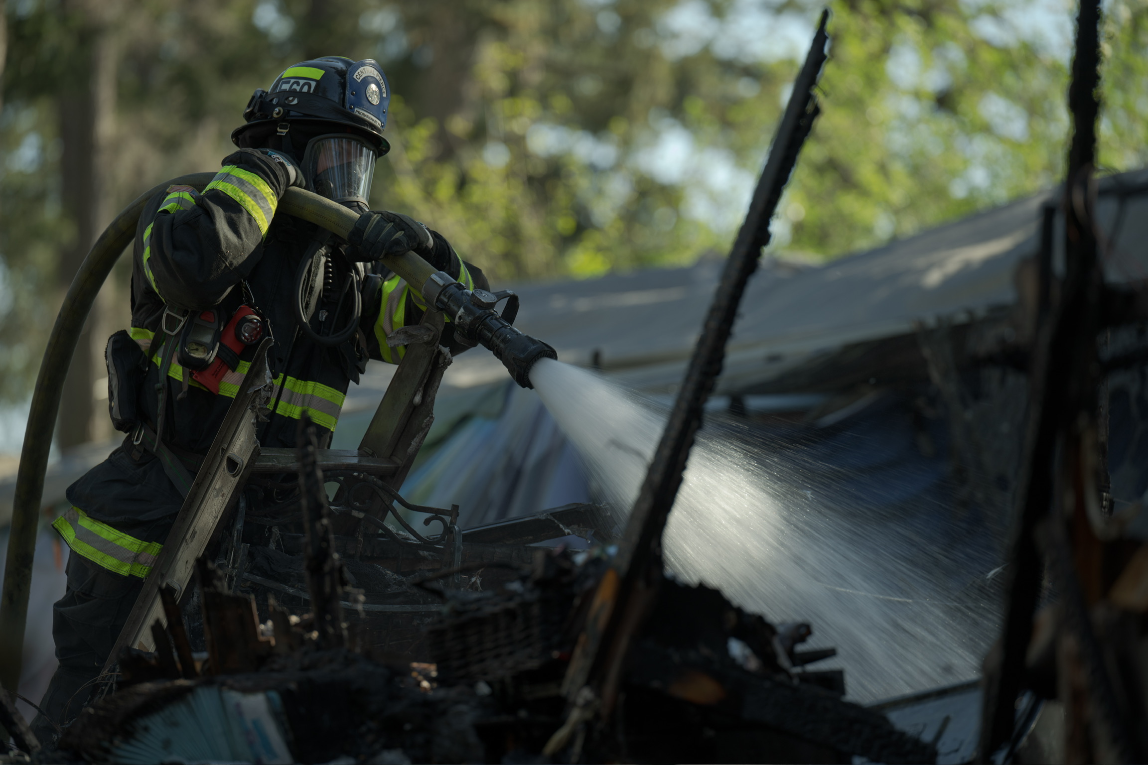A firefighter in full gear sprays water from a hose onto charred debris in an outdoor setting with trees in the background.