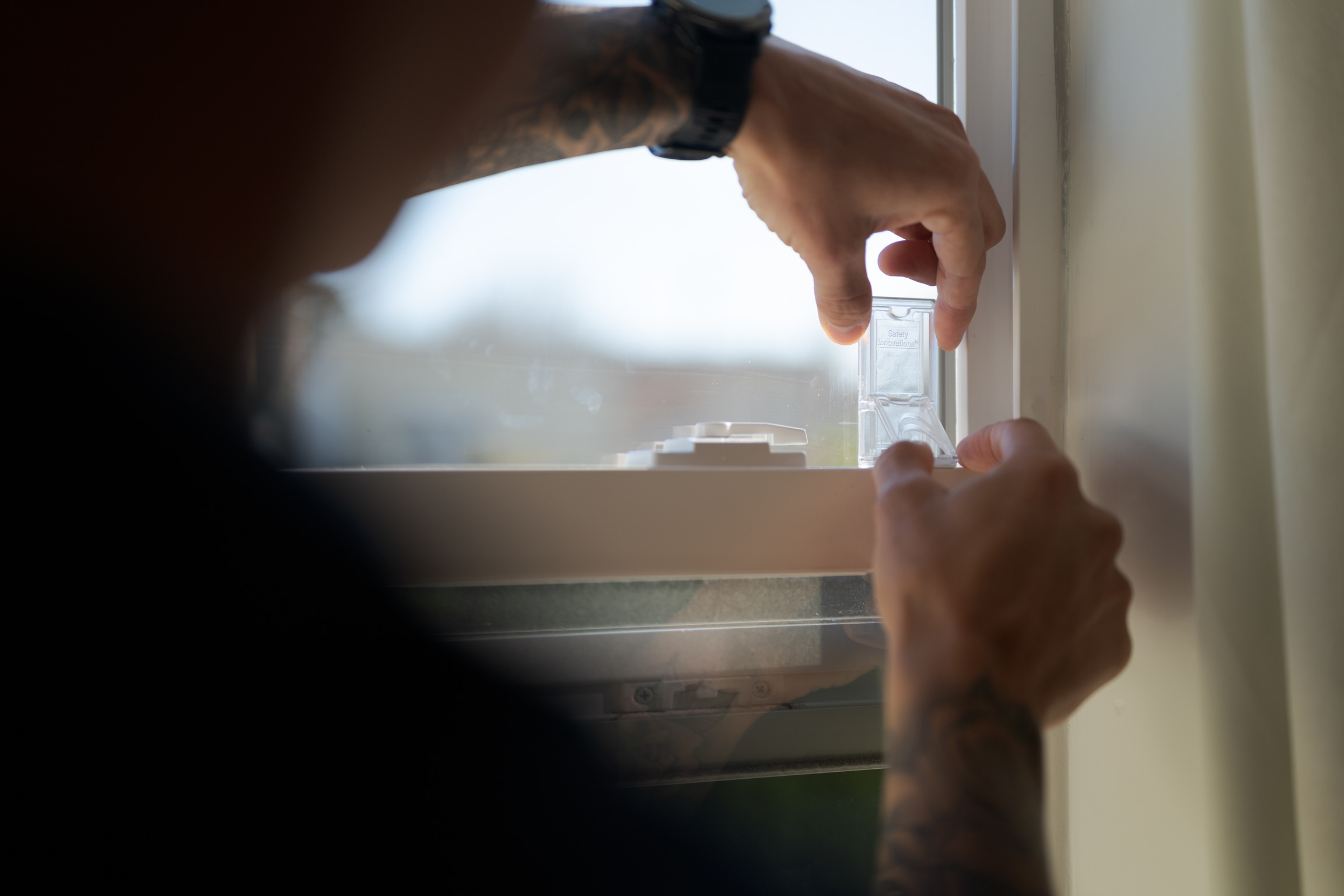 A person with tattoos installs a small clear plastic device on a window frame, with one hand holding the window and the other attaching the device.