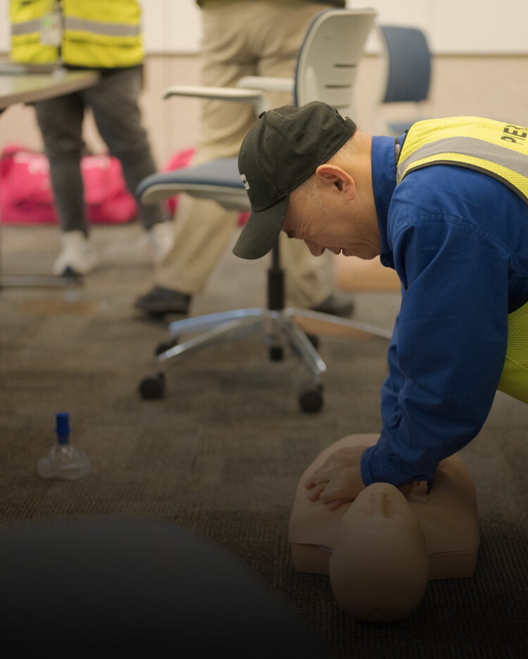 A person wearing a yellow safety vest practices CPR on a mannequin in a training room with chairs and other participants.