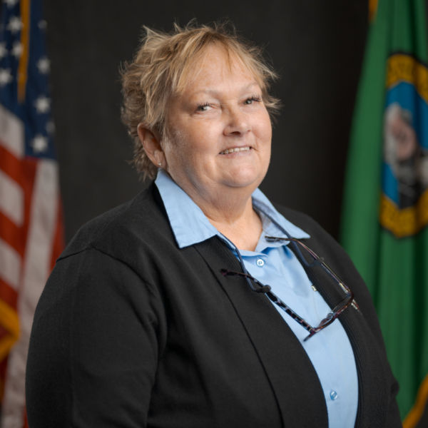 A woman with short blond hair, wearing a blue shirt and black blazer, stands in front of the American and Washington state flags.