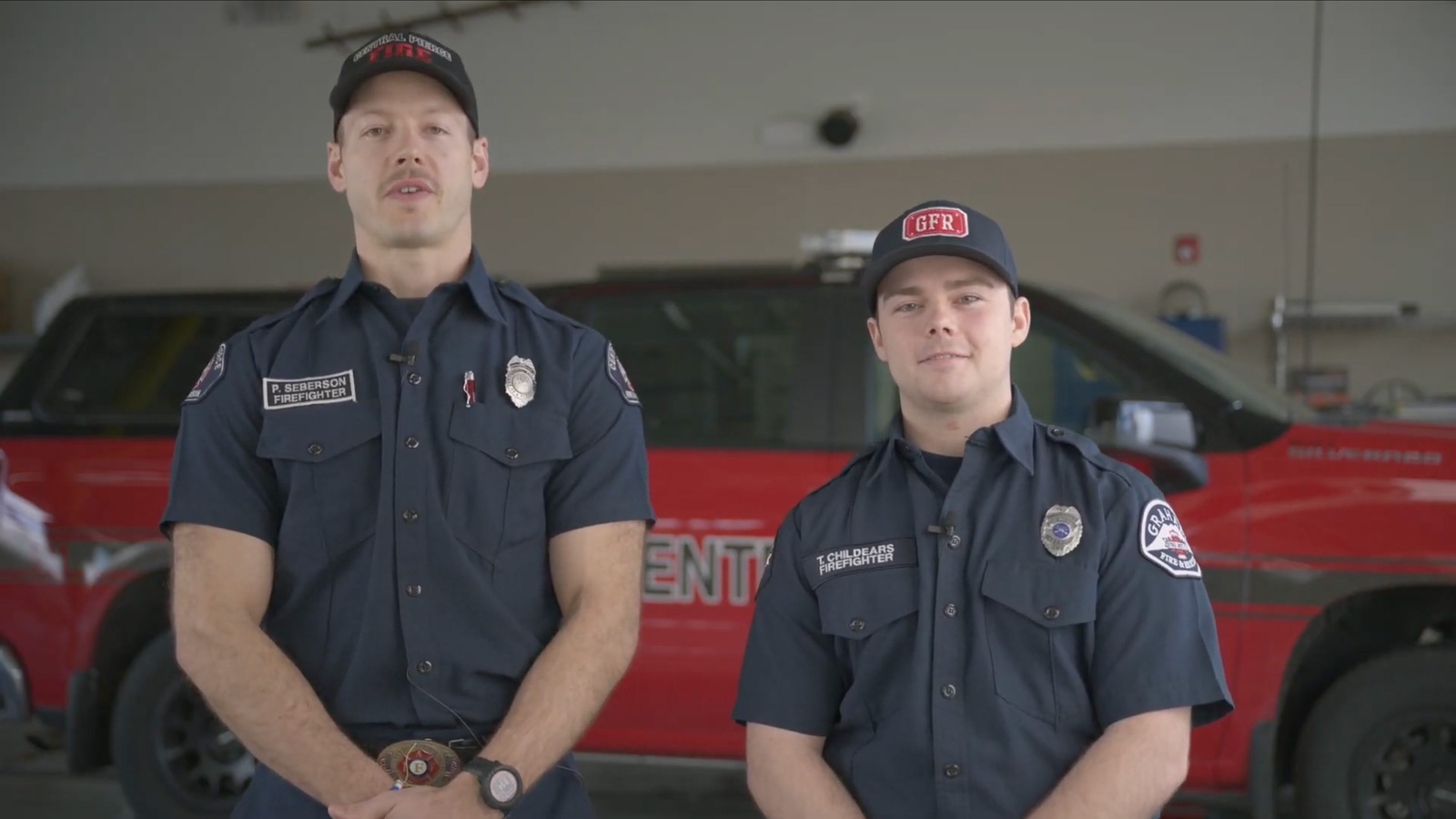 Two firefighters in uniform stand in front of a red emergency vehicle inside a fire station.