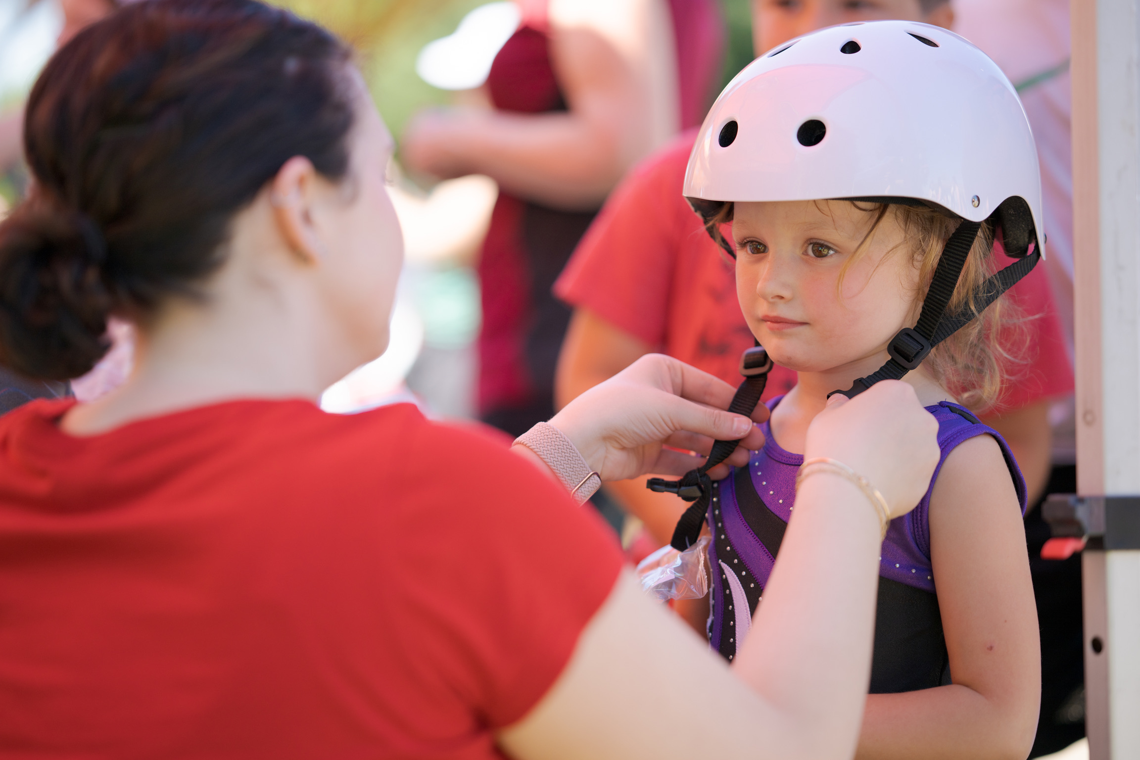 An adult fastens the chin strap of a white helmet on a young girl wearing a purple shirt, with others visible in the background.