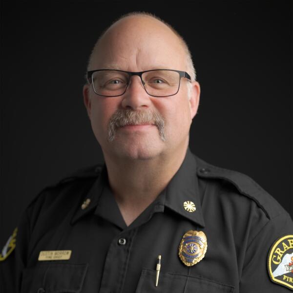 A middle-aged man with glasses and a mustache wearing a black fire department uniform and badges, posing against a dark background.