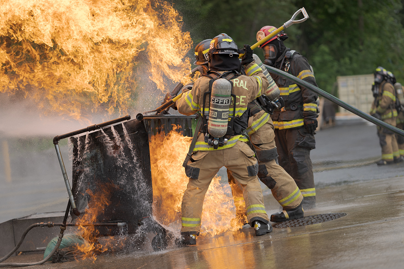 Firefighters in protective gear use hoses to extinguish a large blaze coming from a metal container on wet pavement outdoors, demonstrating skills needed for Central Pierce Fire job opportunities.