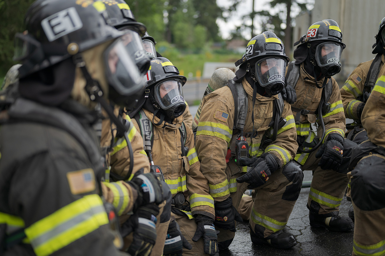 A group of firefighters in full protective gear, including helmets and breathing masks, are kneeling on a wet surface outdoors&mdash;training for Central Pierce Fire job opportunities.