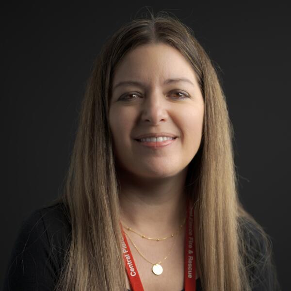 A woman with long straight hair, wearing a black top, gold necklace, and red lanyard, smiles at the camera against a dark background.