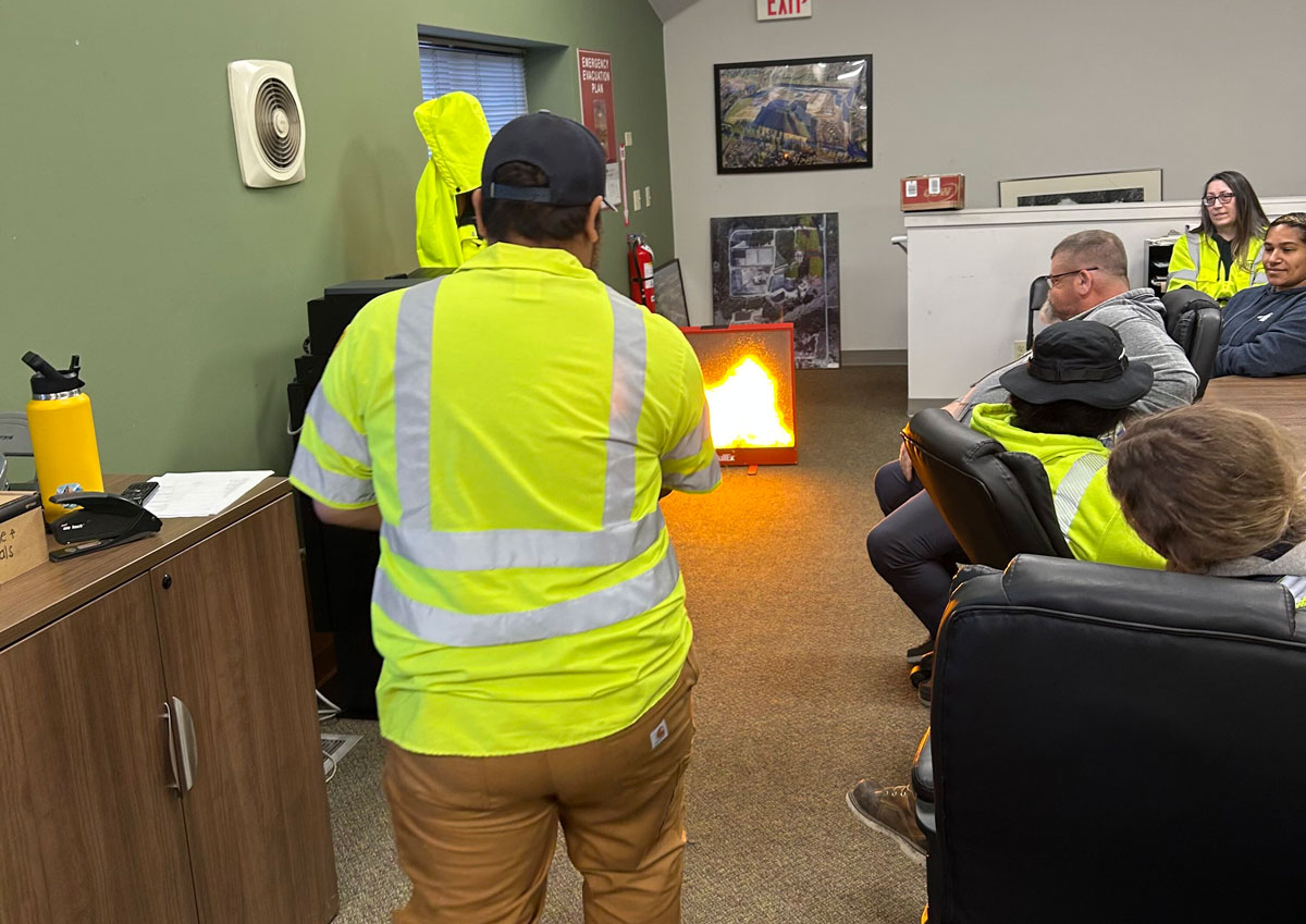 A person in a high-visibility shirt demonstrates fire safety during Central Pierce Fire & Rescue fire extinguisher classes to a seated group, using a small controlled fire in a metal container in the meeting room.