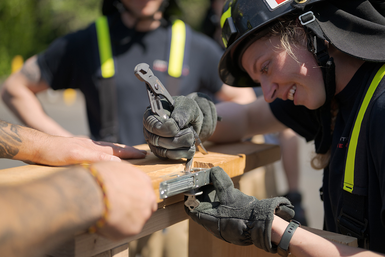 A firefighter in uniform and gloves uses pliers to work on a wooden structure while others assist nearby&mdash;showcasing the teamwork valued in Central Pierce Fire & Rescue becoming a firefighter.