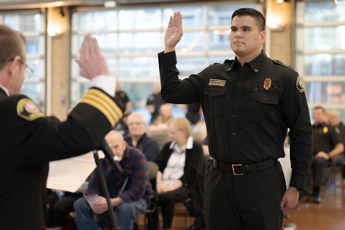 A uniformed man raises his right hand while being sworn in at a formal ceremony, with seated attendees visible in the background.