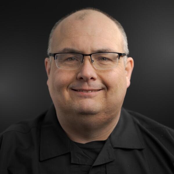 A middle-aged man with glasses and a black shirt smiles at the camera against a plain dark background.