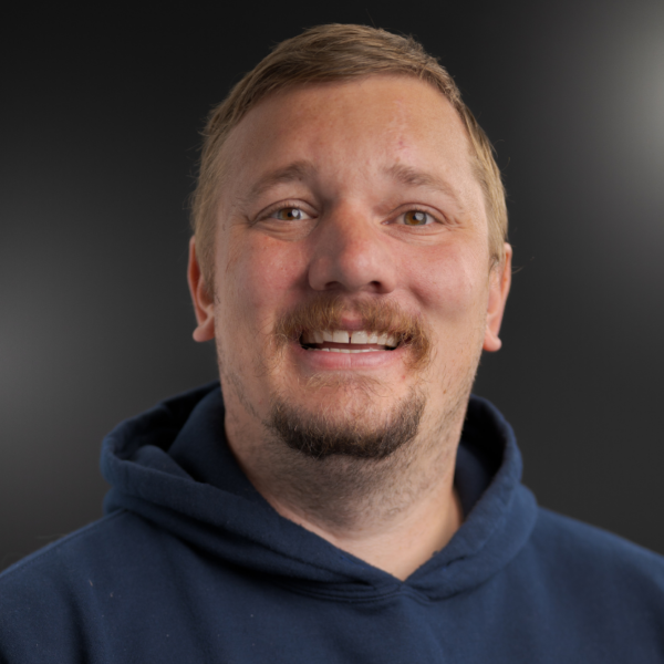 A man with short light brown hair and facial hair is smiling, wearing a navy hoodie, posed against a dark, blurred background.