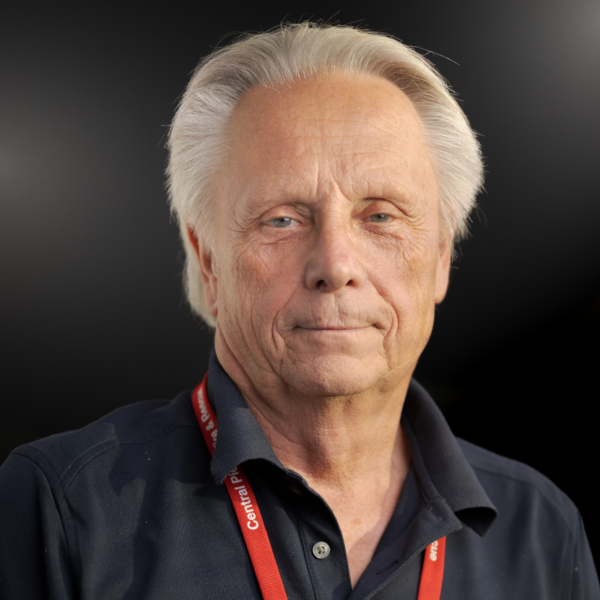 An older man with gray hair wearing a dark shirt and a red lanyard stands against a dark background, looking directly at the camera with a neutral expression.