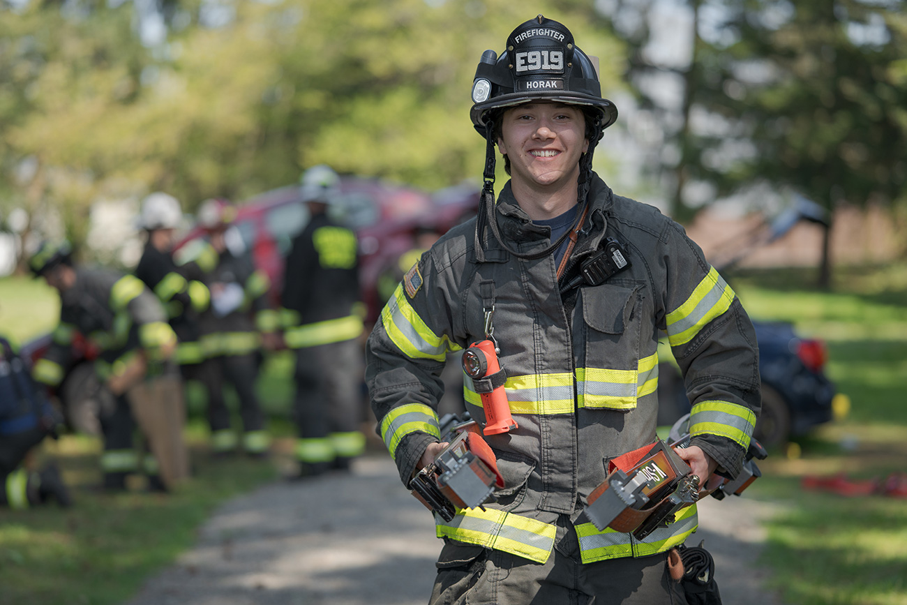 A firefighter in uniform stands smiling outdoors, holding equipment, with other firefighters and an emergency scene blurred in the background&mdash;an inspiring look at how to become a firefighter at Central Pierce Fire & Rescue.