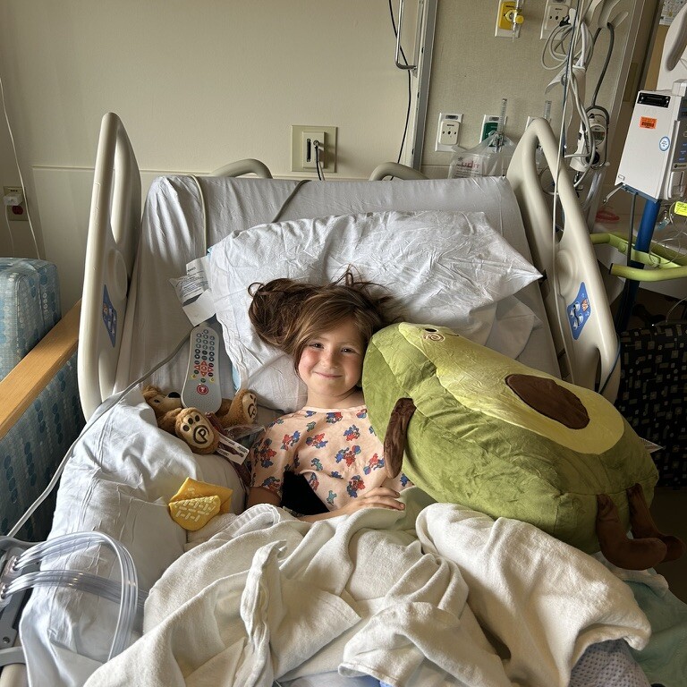 A young child is lying in a hospital bed, smiling, surrounded by stuffed animals and blankets, with medical equipment visible in the background.