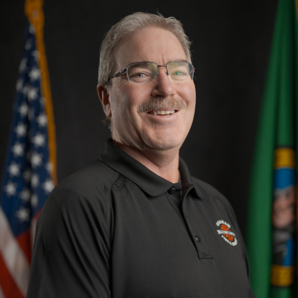 A man with gray hair and glasses, wearing a black polo shirt, stands smiling in front of the U.S. and Washington state flags.