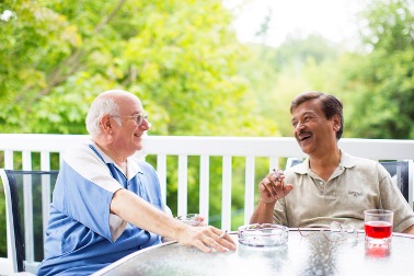 Two men sit at an outdoor table, smiling and talking over drinks and an ashtray. Green trees are visible in the background, highlighting a relaxed setting that underscores the importance of CPFR safety for older adults.