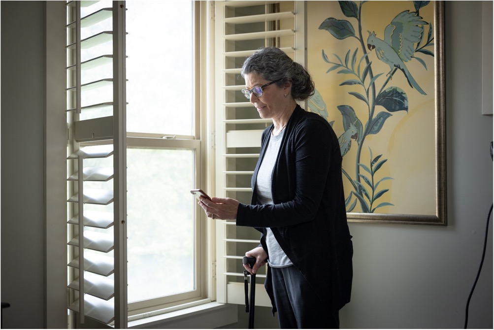 An older woman with gray hair stands by a window holding a cane and a smartphone, wearing glasses and a black cardigan. A bird painting hangs on the wall beside her&mdash;a scene reflecting the importance of fall prevention for older adults.