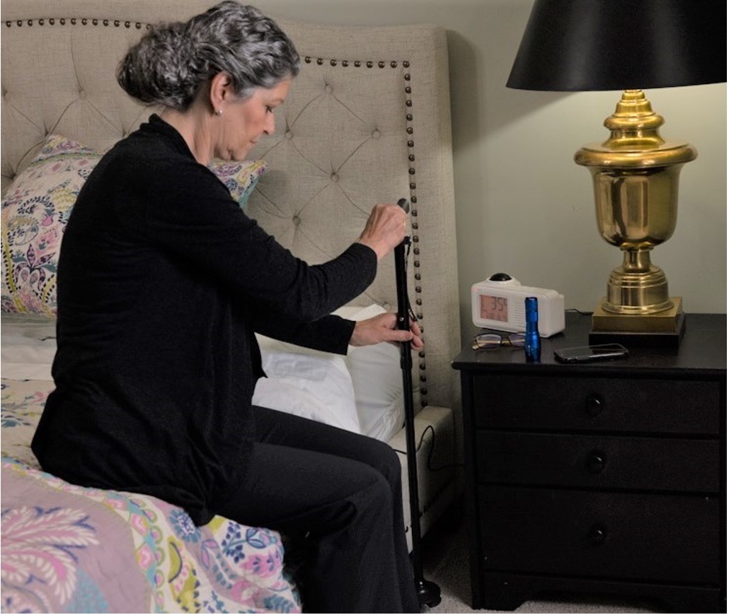 A woman sits on the edge of a bed, holding a cane beside a nightstand with a lamp, alarm clock, flashlight, and phone&mdash;emphasizing fall prevention for older adults and safe nighttime routines.